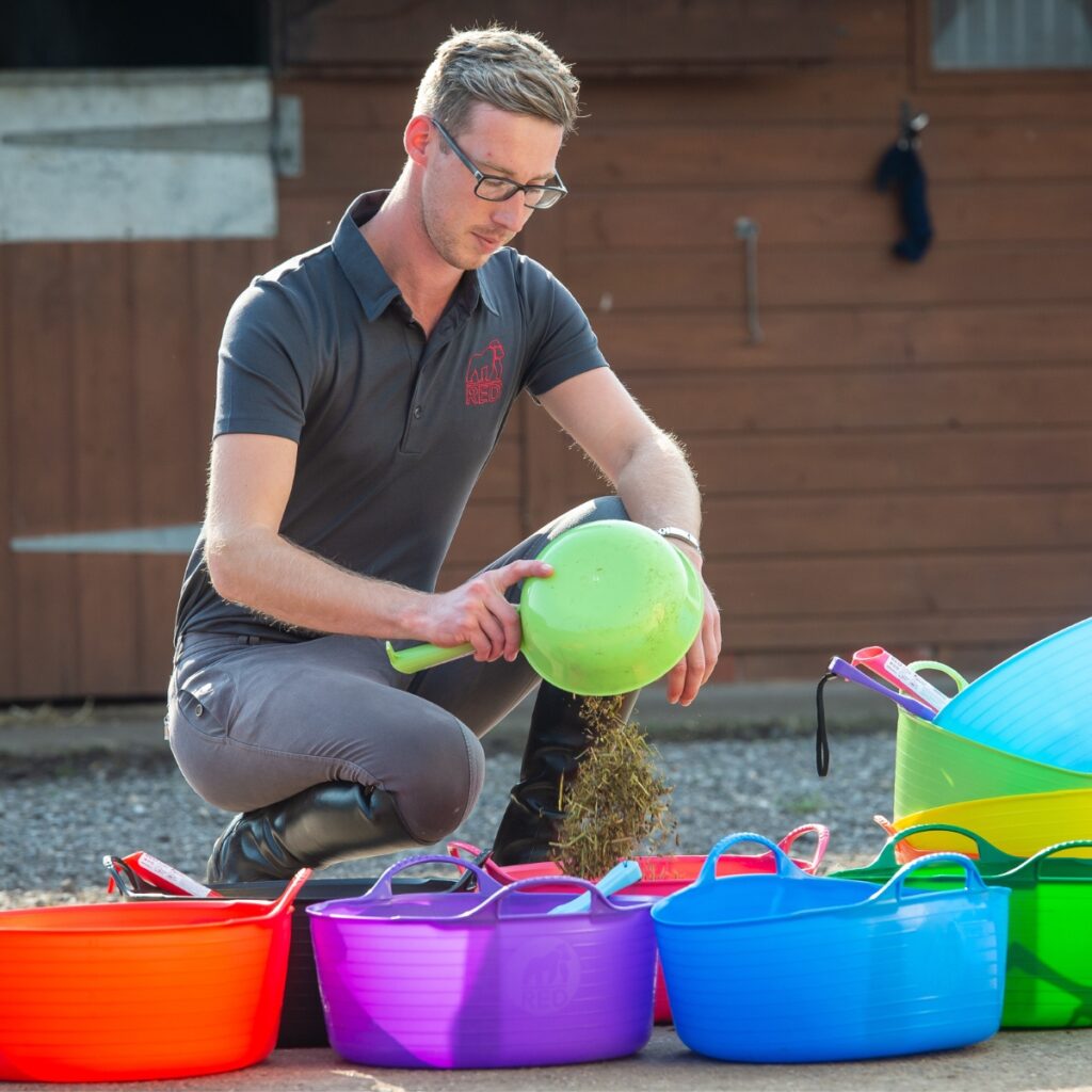 Man using a feed scoop to measure out and prepare horse feed in a range of feed buckets.